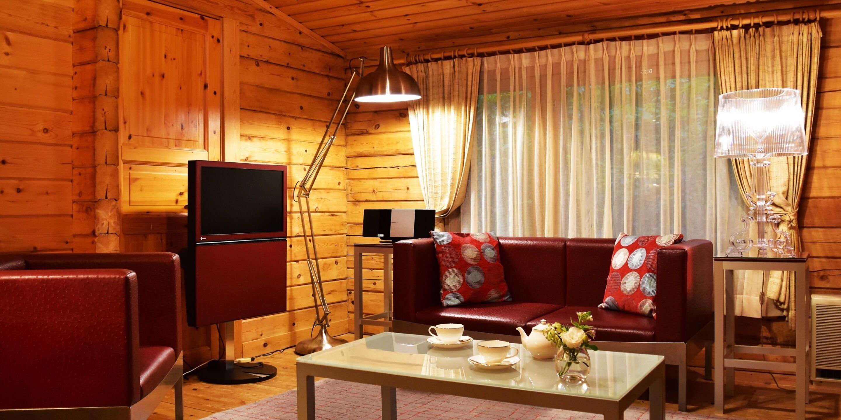 Une pièce chaleureuse avec des murs en bois, un canapé rouge, une télévision, une table avec un service à thé et une lampe.