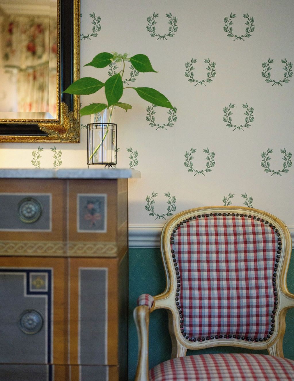 A room with a vintage chair, a mirror, and a wall with a pattern of laurel wreaths.