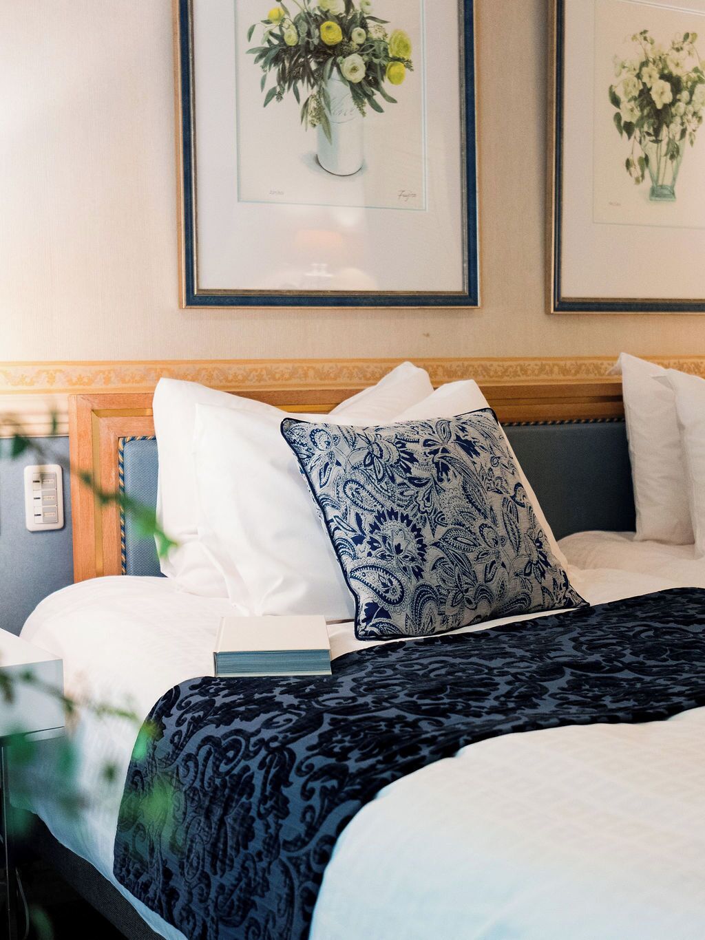 Un lit bien fait, agrémenté d’oreillers blancs et bleus à motifs, d’une couvre-lit bleu foncé et de deux tableaux floraux encadrés accrochés au mur.