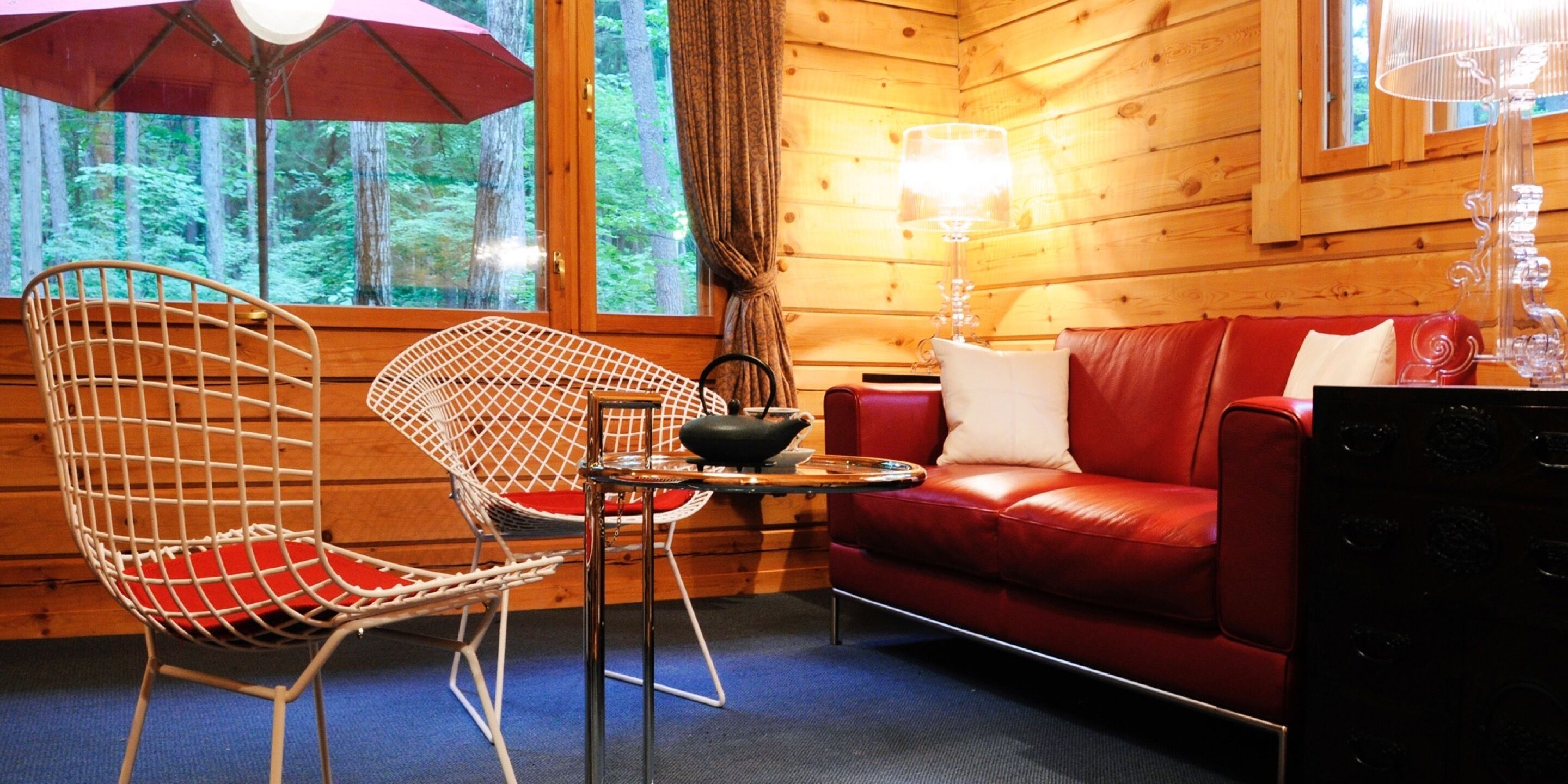 A cozy cabin interior with a red leather sofa, a white wire chair, a small table with a lamp, and a view of the forest through large windows.