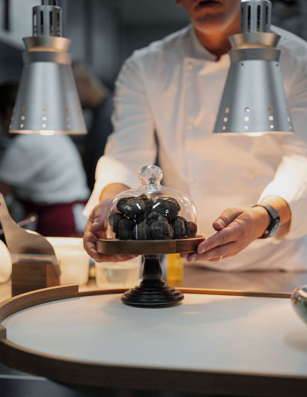 A chef holding a dish with a glass dome over it, standing in a kitchen with two industrial kitchen lights.