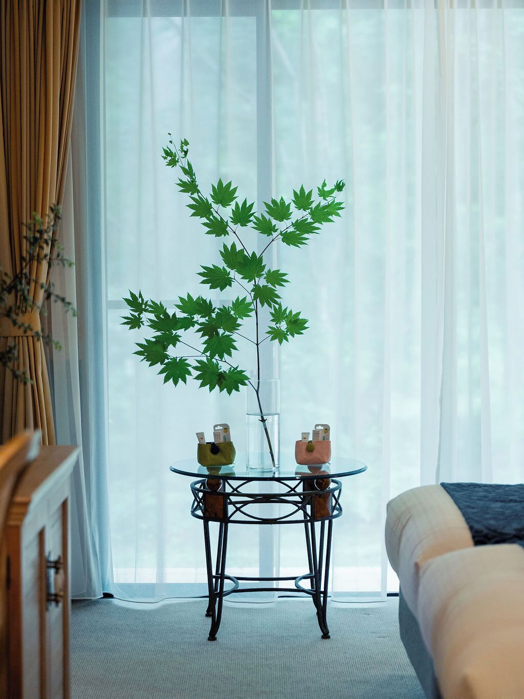 Una habitación con una cama, un ventanal con cortinas translúcidas, una mesa con dos recipientes de vidrio y una planta verde grande.