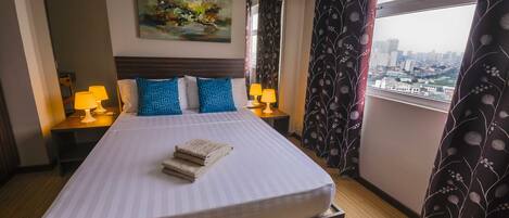A hotel room with a large bed, two bedside lamps, a painting on the wall, and a view of a cityscape through the window.