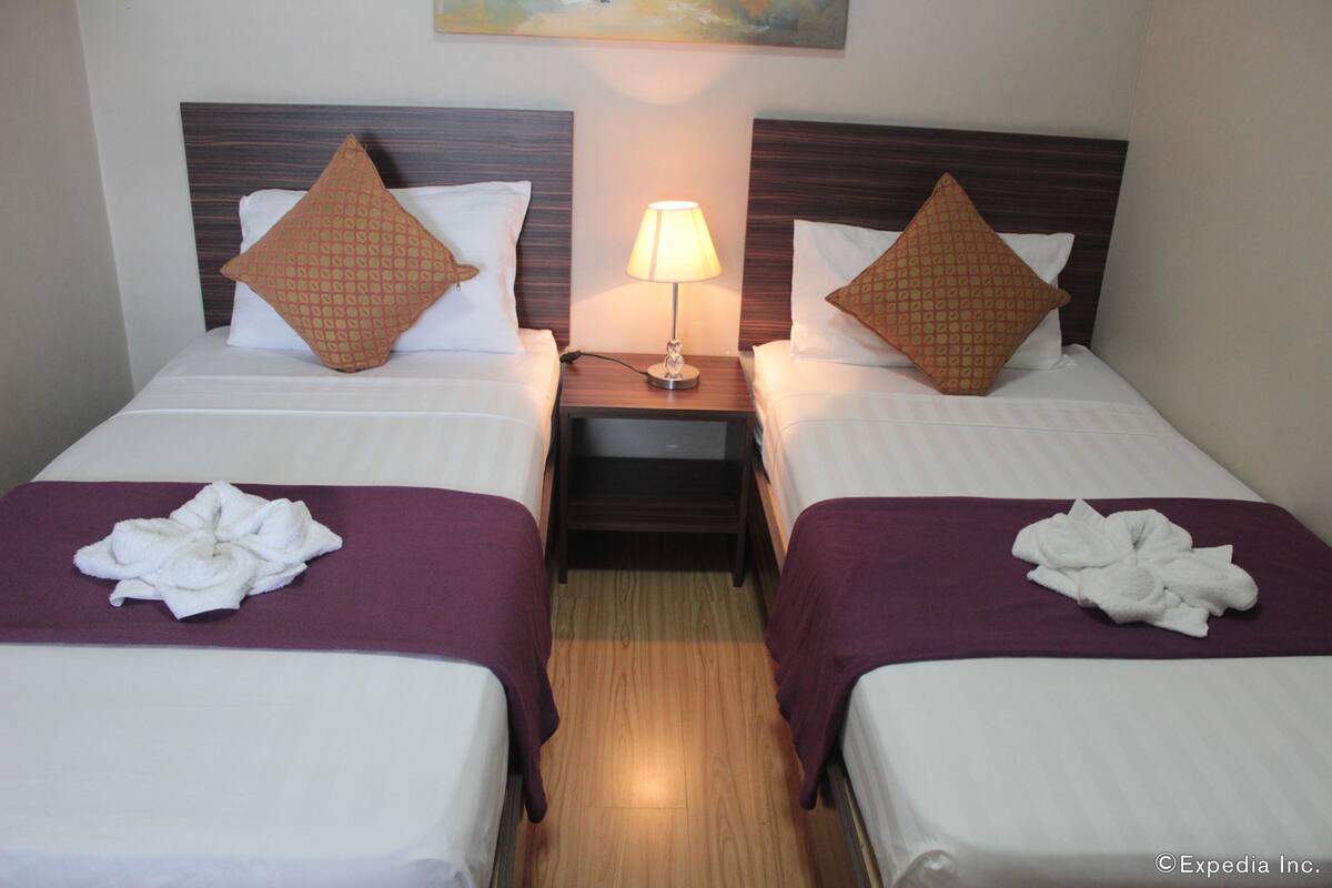 A hotel room with two beds, each with a wooden headboard, a nightstand with a lamp, and towels folded on the beds.