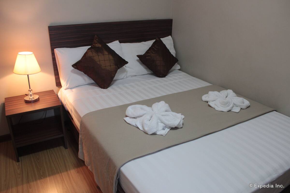 A neatly made bed with white linens and two brown decorative pillows. A white towel is placed on the bed, and a lamp is on the nightstand beside the bed.