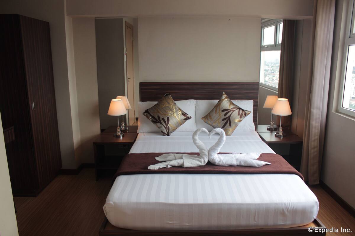 A hotel room with a large bed, two nightstands with lamps, a wooden wardrobe, and a view through the window.