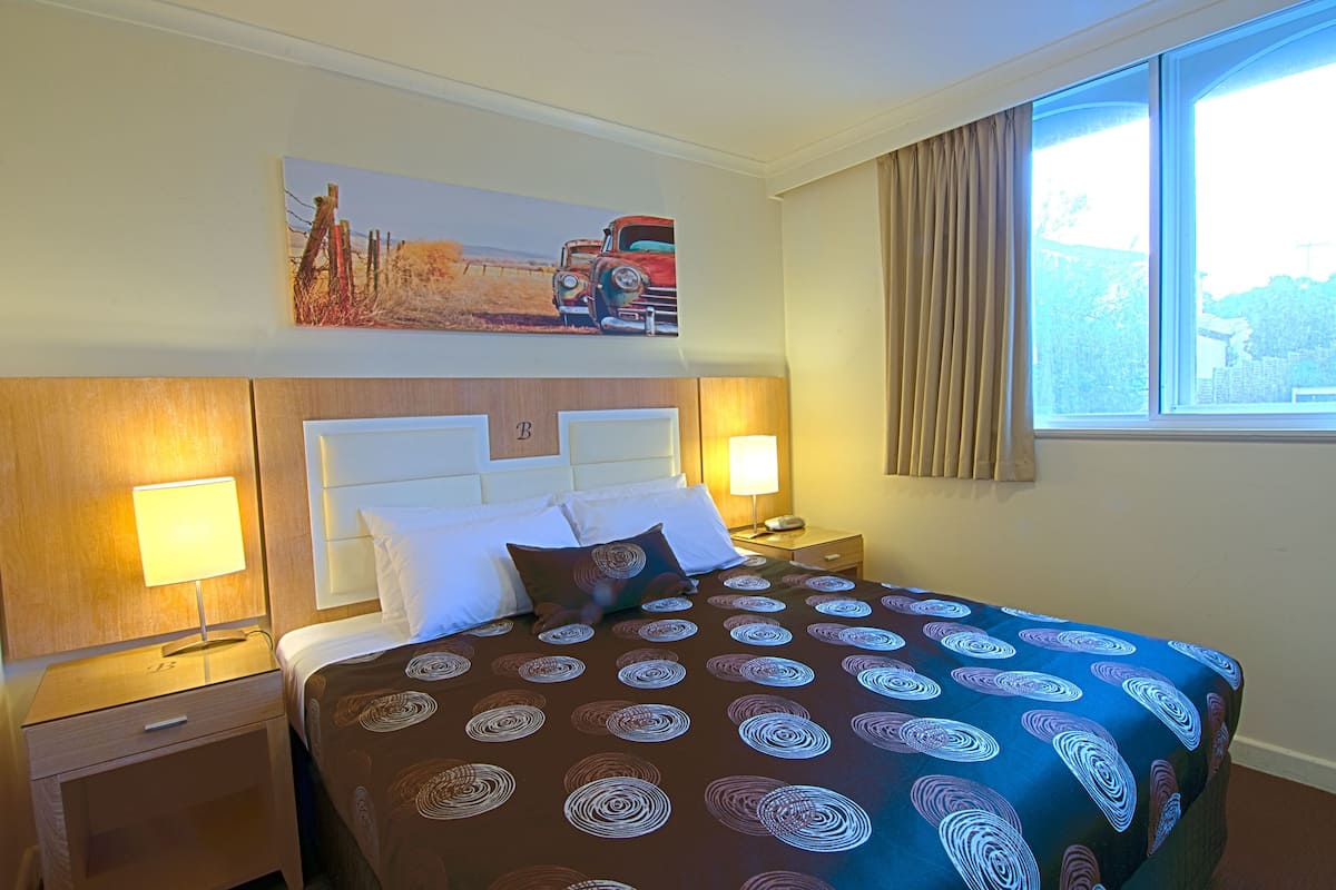 Una habitación de hotel con cama, dos mesitas de noche, lámpara, un cuadro en la pared y ventana con cortinas.