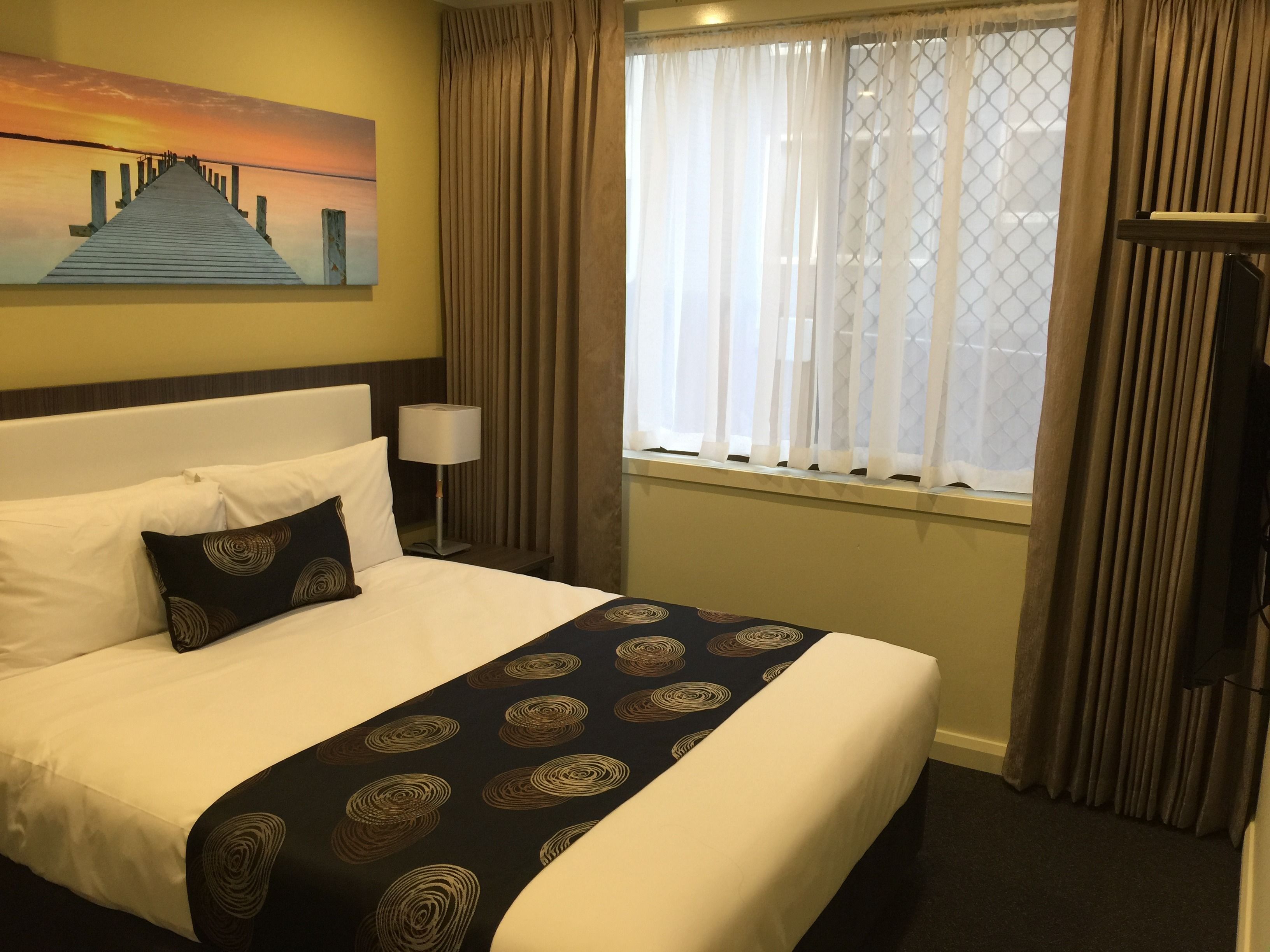 Una camera d'hotel con un letto, un quadro raffigurante un tramonto su un molo, una finestra con tende e un comodino con una lampada.
