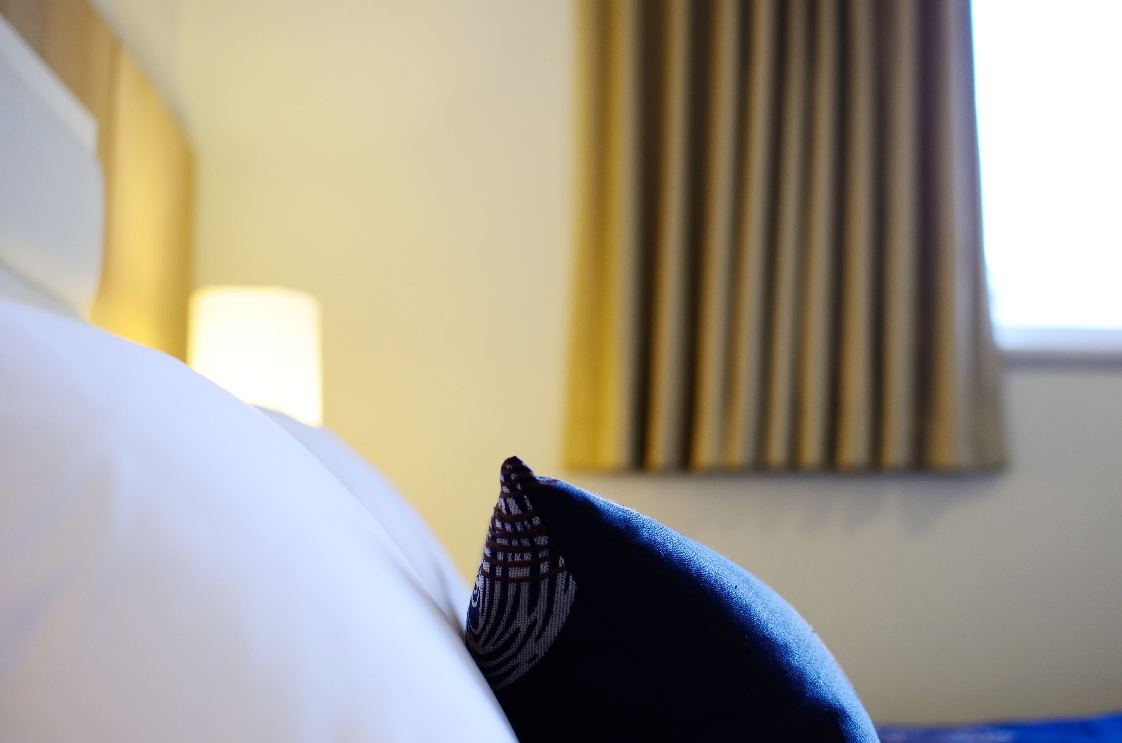 A close-up of a bed with a white pillow and a patterned pillow, a lamp in the background, and a window with blinds.