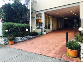 Entrada de um edifício com um caminho pavimentado que leva a uma porta de vidro. Há plantas em vasos e uma parede de pedra à esquerda.