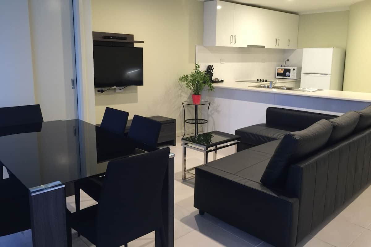 Una sala de estar moderna con un sofá negro, una mesa de comedor con sillas, un televisor de pantalla plana montado en la pared y una zona de cocina con armarios y electrodomésticos.