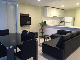 Uma sala de estar moderna com sofá preto, mesa de jantar com cadeiras, televisão de tela plana na parede e cozinha com armários e eletrodomésticos.