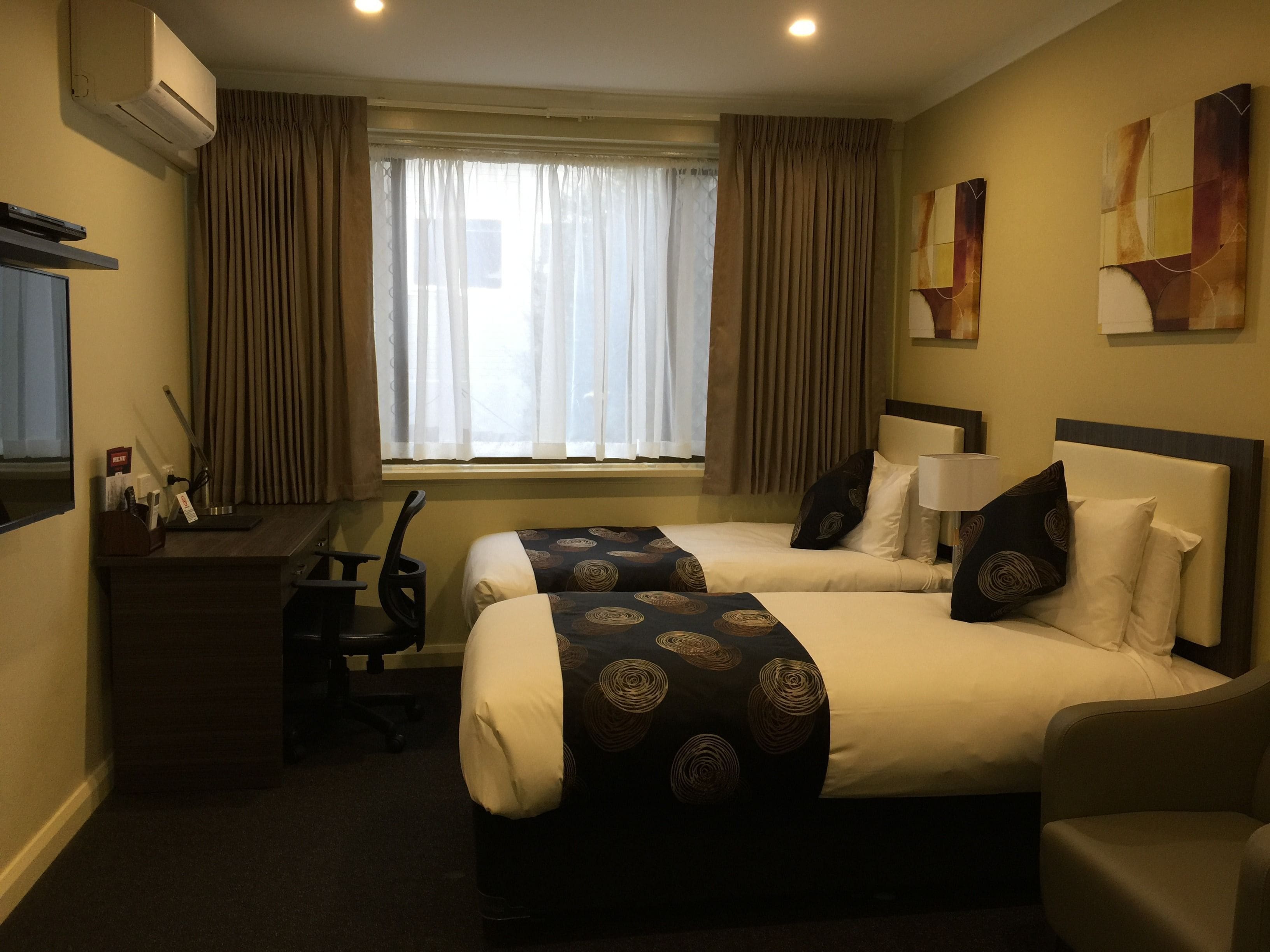 A hotel room with two beds, a desk, a chair, a TV, and a window with curtains.