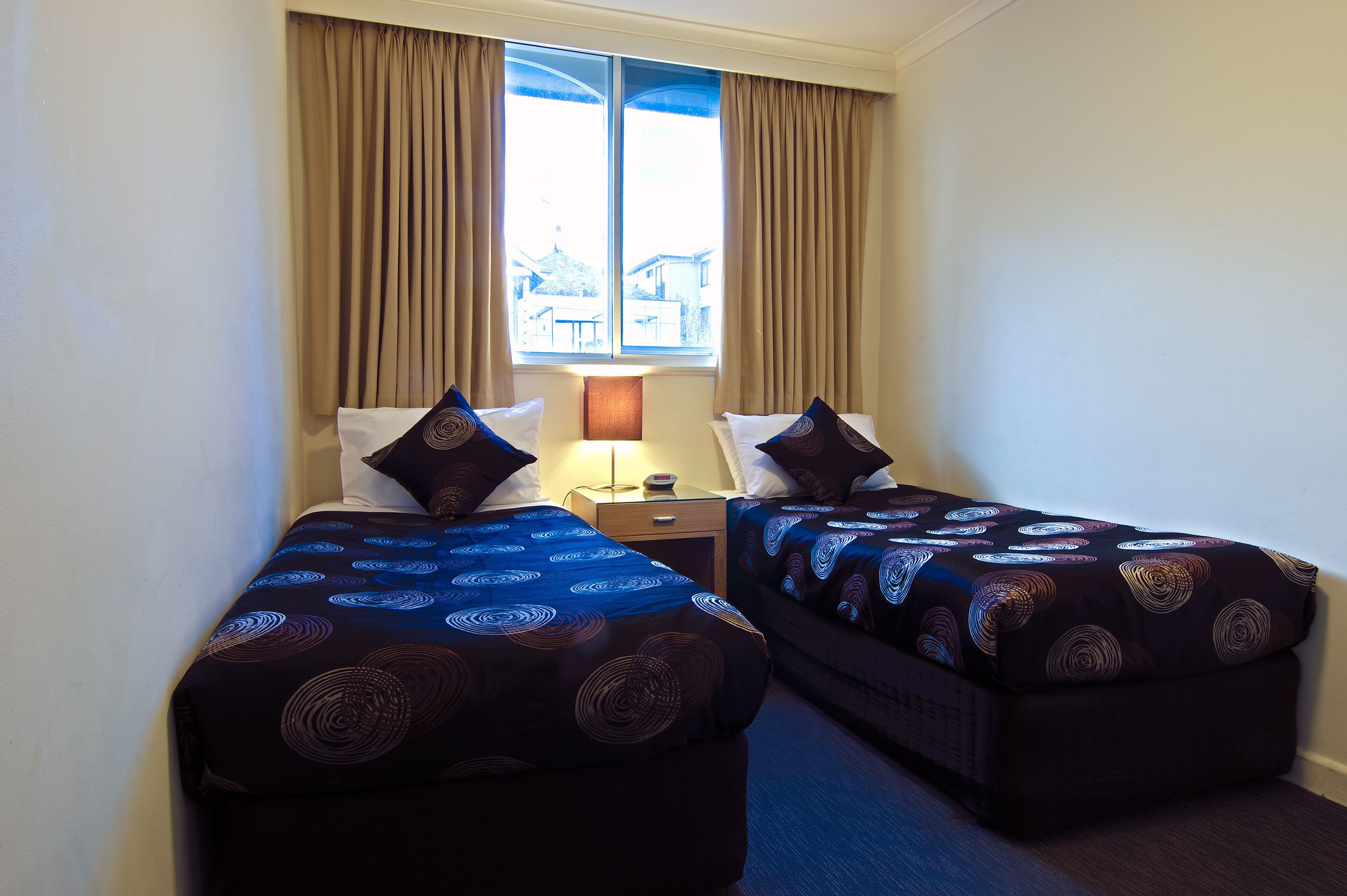 A hotel room with two beds, a nightstand, a lamp, and a window with curtains.