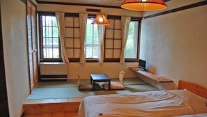 Ein Zimmer mit einem Bett, einem kleinen Tisch und einer Bank. Es gibt Fenster mit Vorhängen und eine Hängelampe.