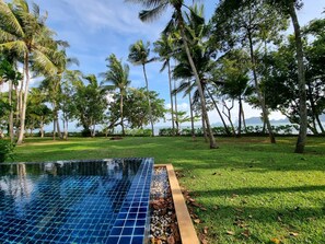 A swimming pool with clear blue tiles, surrounded by a grassy area and palm trees, with a view of the sea and mountains in the background. - Amatapura Beach Villa 12 (Krabi)