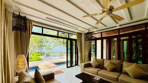 A spacious living room with a large sofa, a coffee table, and a ceiling fan. There is a large window providing a view of a pool and greenery outside.