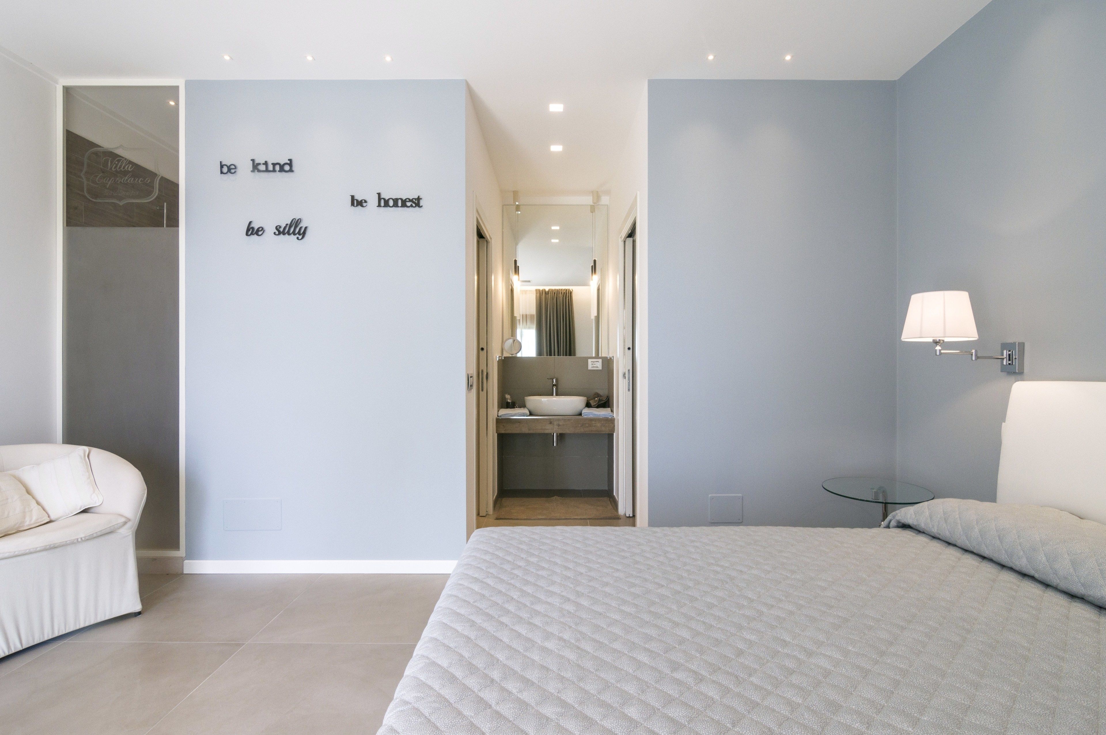 A modern bedroom with a bed, a white chair, a bathroom with a sink and mirror, and motivational wall art.