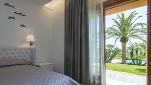 A bedroom with a bed, a nightstand, a lamp, a window with curtains, and a view of a pool and palm trees.