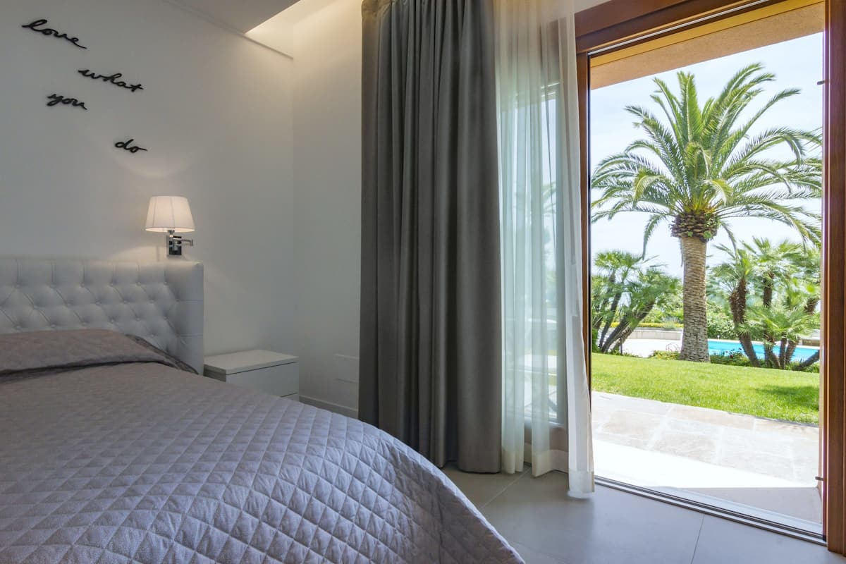 A bedroom with a bed, a nightstand, a lamp, a window with curtains, and a view of a pool and palm trees.