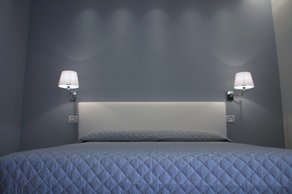 Una cama con una colcha azul acolchada, dos lámparas de pared y un cabecero blanco.