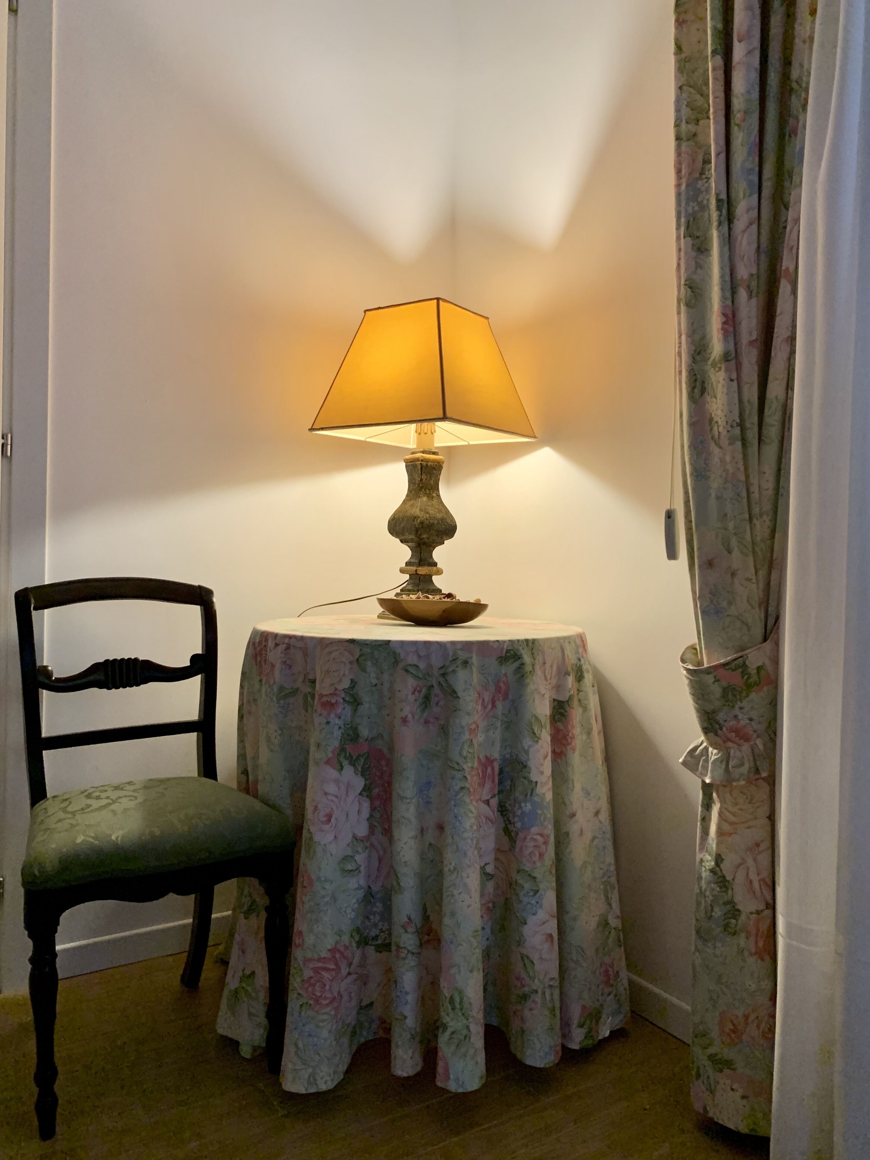 Een kamer met een tafel, overdekt met een bloemendessin, een stoel, een lamp en een gordijn.