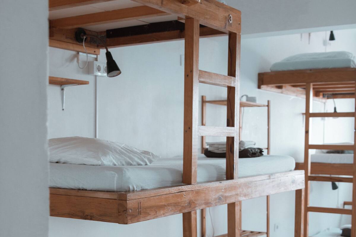 Camas literas con estructuras de madera y ropa blanca, dispuestas en una habitación tipo dormitorio con paredes blancas y suelo de baldosas.