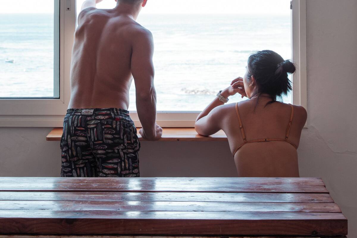 Una habitación con un gran ventanal con vista al mar, un banco de madera y una persona de pie junto a la ventana.