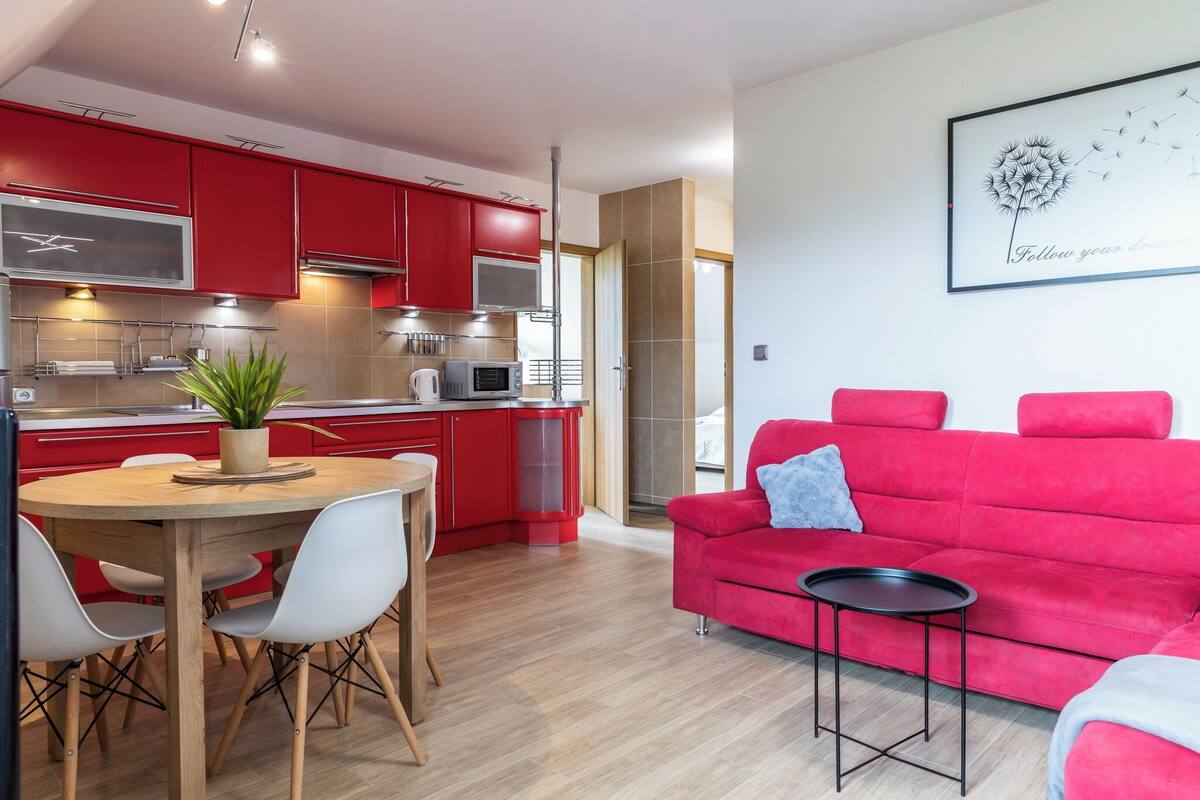 A modern kitchen with red cabinets, a dining table with chairs, a red sofa, and a framed artwork on the wall.
