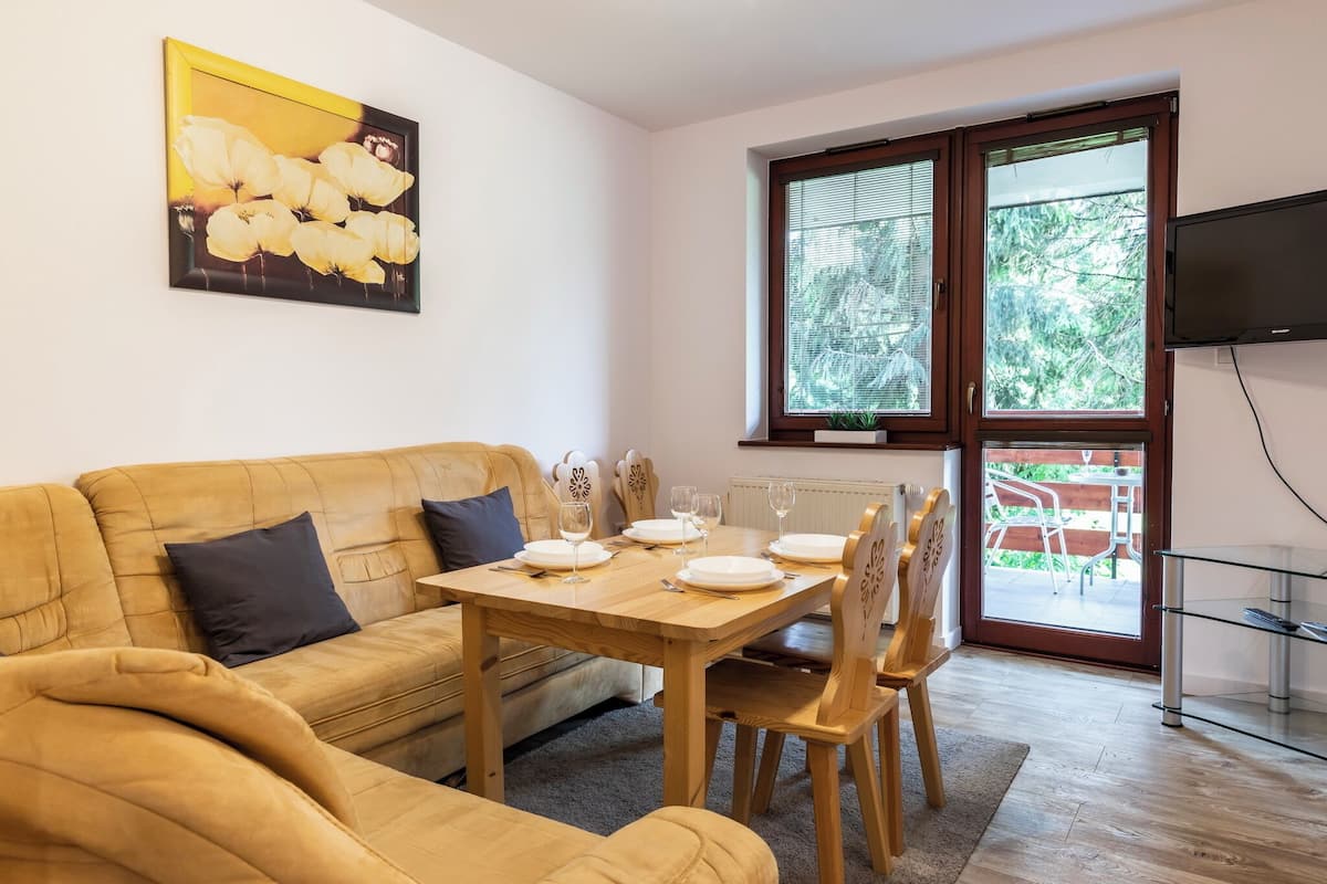 A living room with a sofa, a dining table set with plates and glasses, a TV mounted on the wall, and a window with a view of the outdoors.