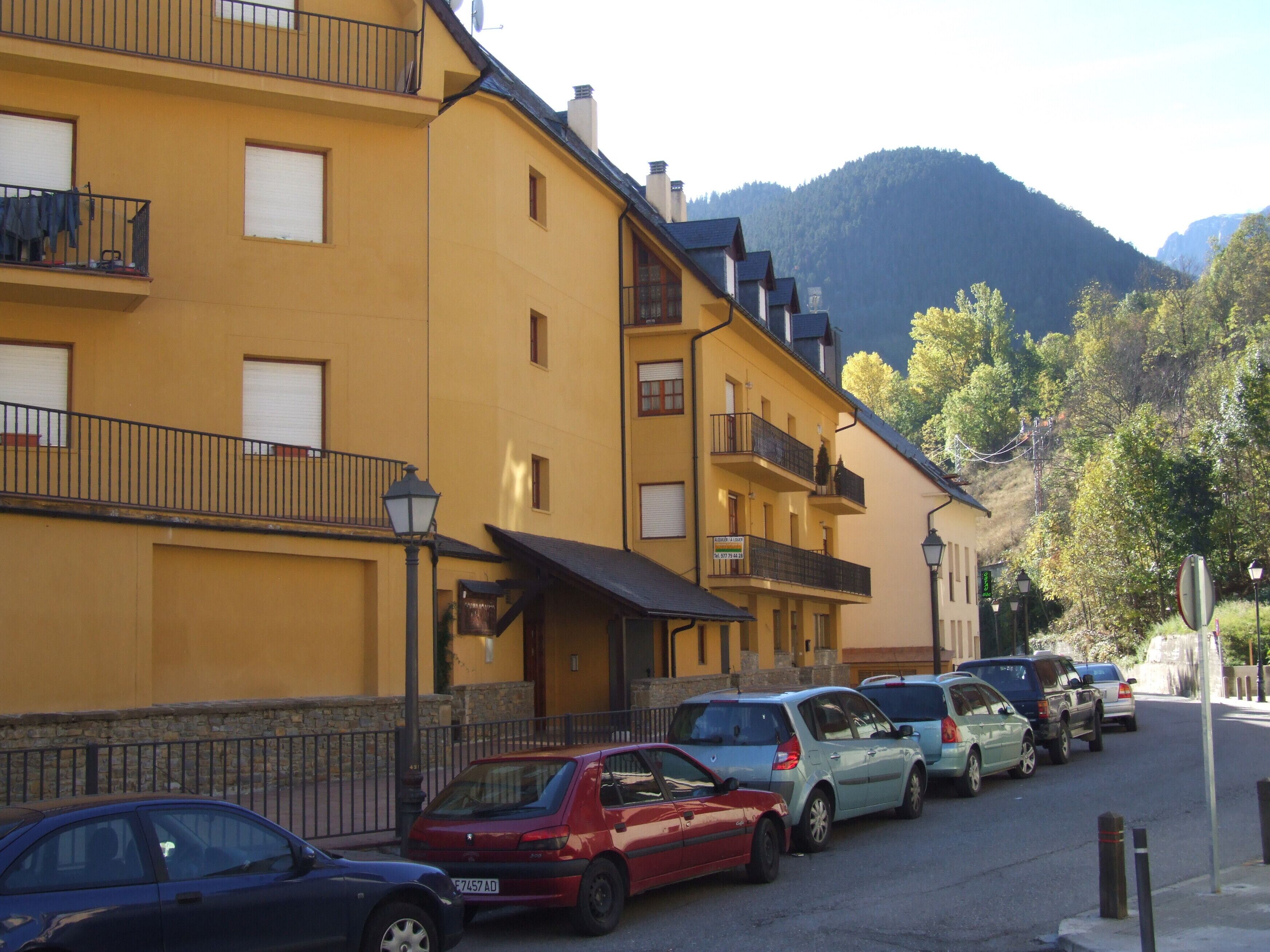 Fila de edificios amarillos con balcones, coches aparcados y montañas al fondo.