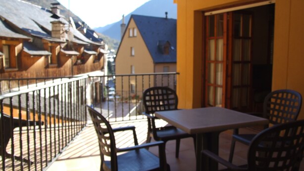A balcony with a table and chairs, overlooking a mountain and a street with buildings.