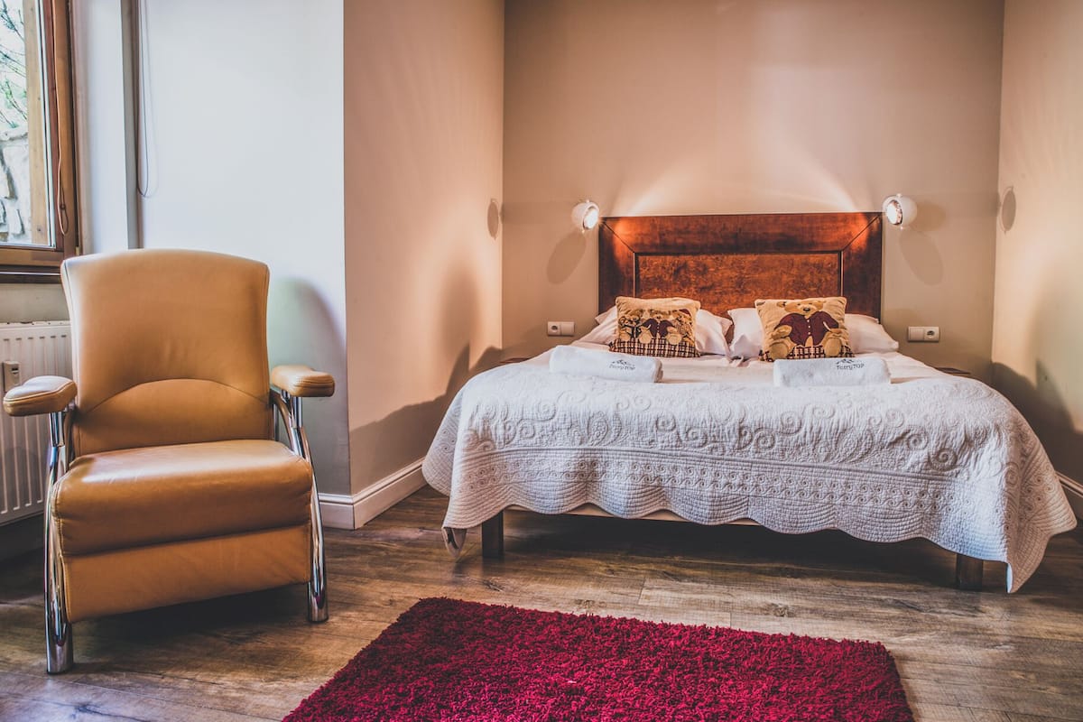 Un dormitorio con una cama, una silla, una alfombra roja y piso de madera.