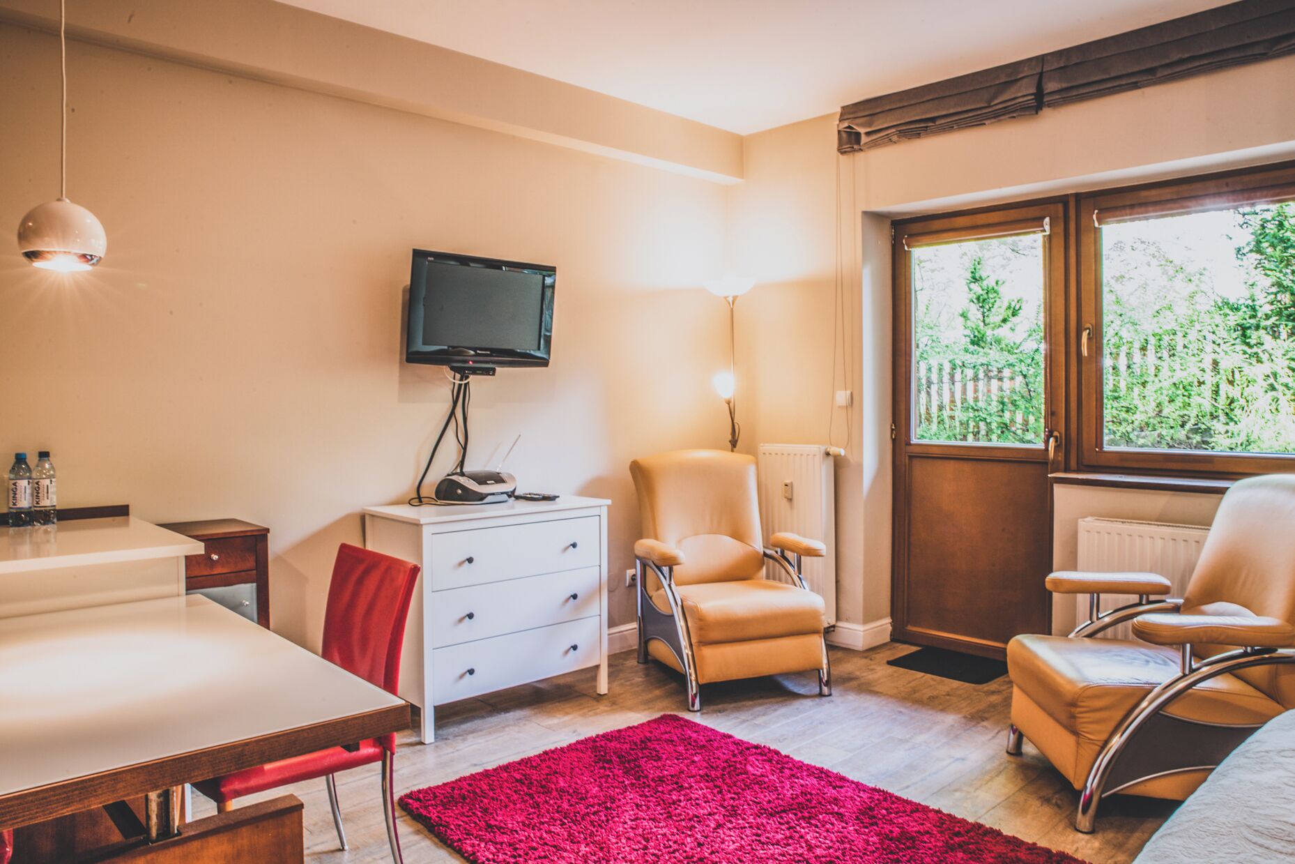 A room with a TV, a desk, two chairs, a red rug, and a window with a view of greenery.