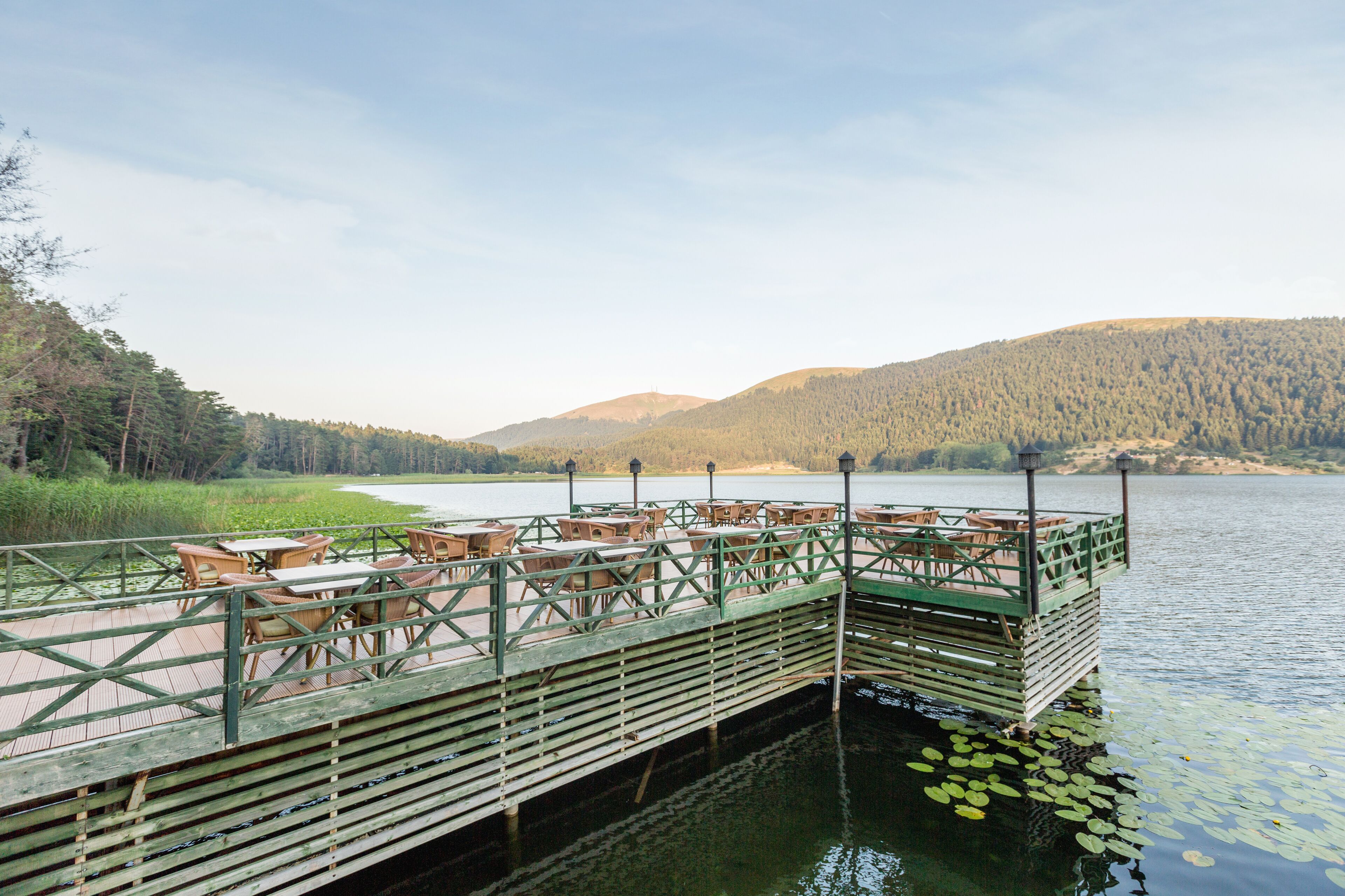 Un muelle de madera con mesas y sillas se extiende sobre un lago, rodeado de colinas cubiertas de bosque.