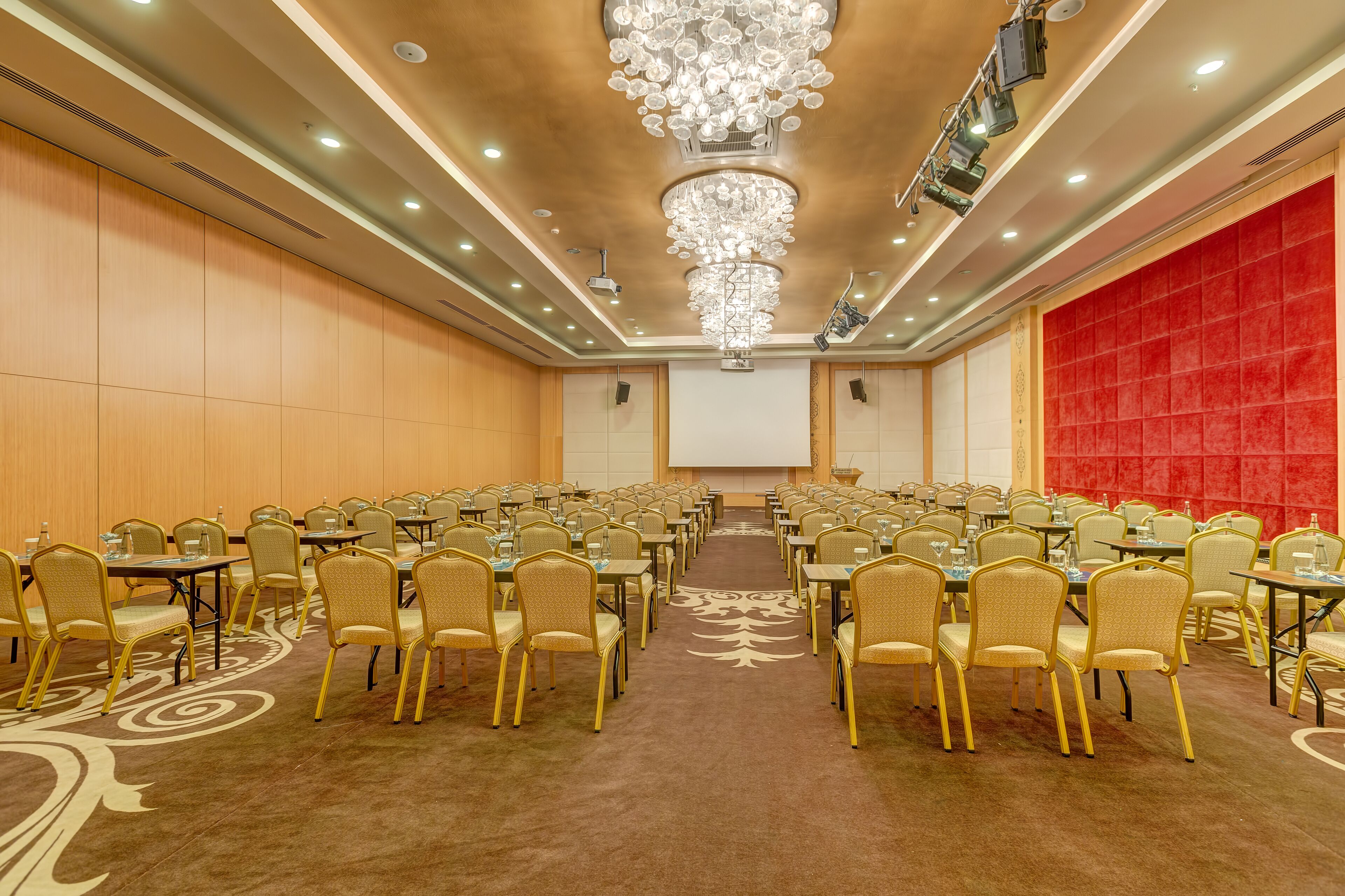 Une salle de conférence avec des rangées de tables et de chaises, un grand lustre et un mur d’accent rouge.