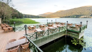 Terraza de madera con mesas y sillas que da a un lago, rodeada de vegetación y colinas.