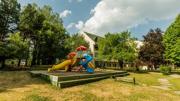 A playground with slides and climbing structures, surrounded by trees and a building in the background.