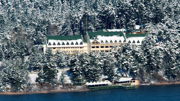Un edificio grande de varios pisos con techo verde, rodeado de árboles cubiertos de nieve y una masa de agua.