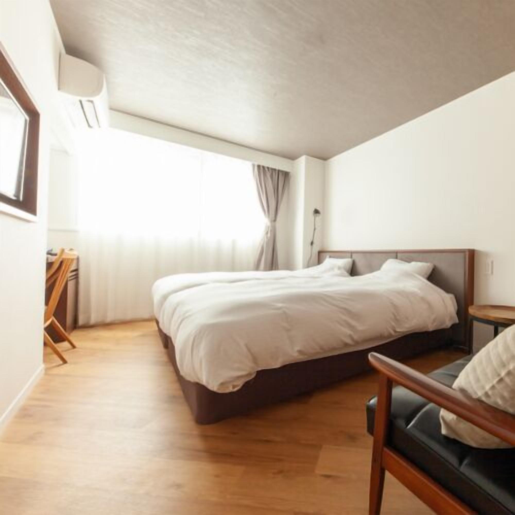 A bedroom with a double bed, a bedside table, a wooden floor, and a window with curtains.