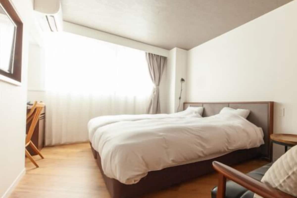 A bedroom with a double bed, a bedside table, a wooden floor, and a window with curtains.