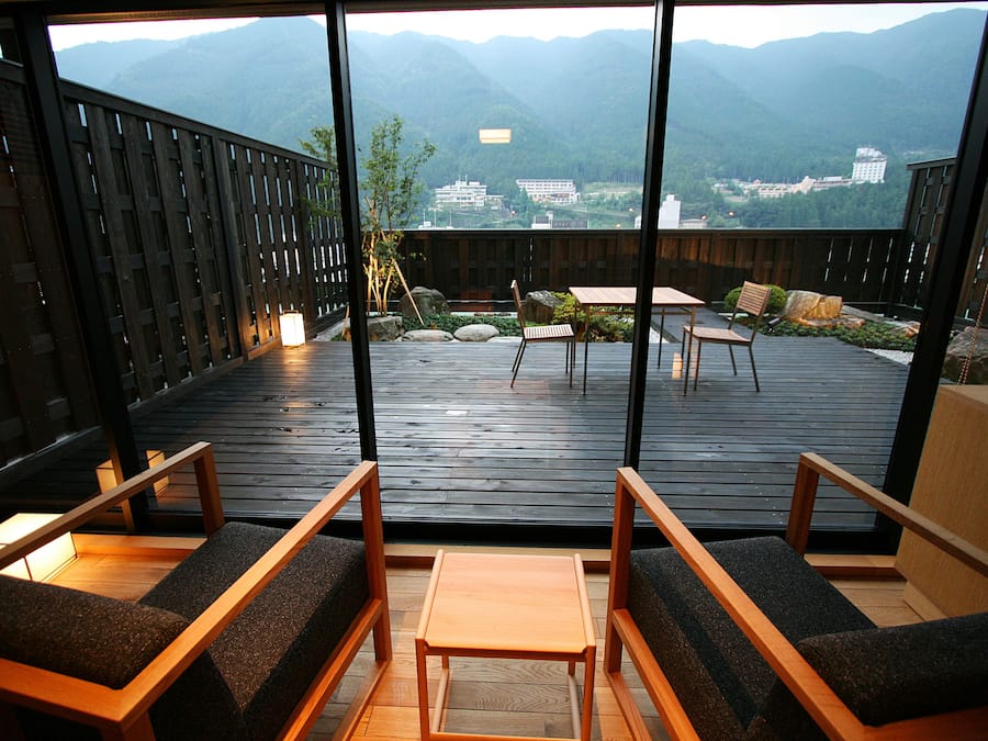 A wooden deck with seating and a table, offering a view of mountains and buildings.