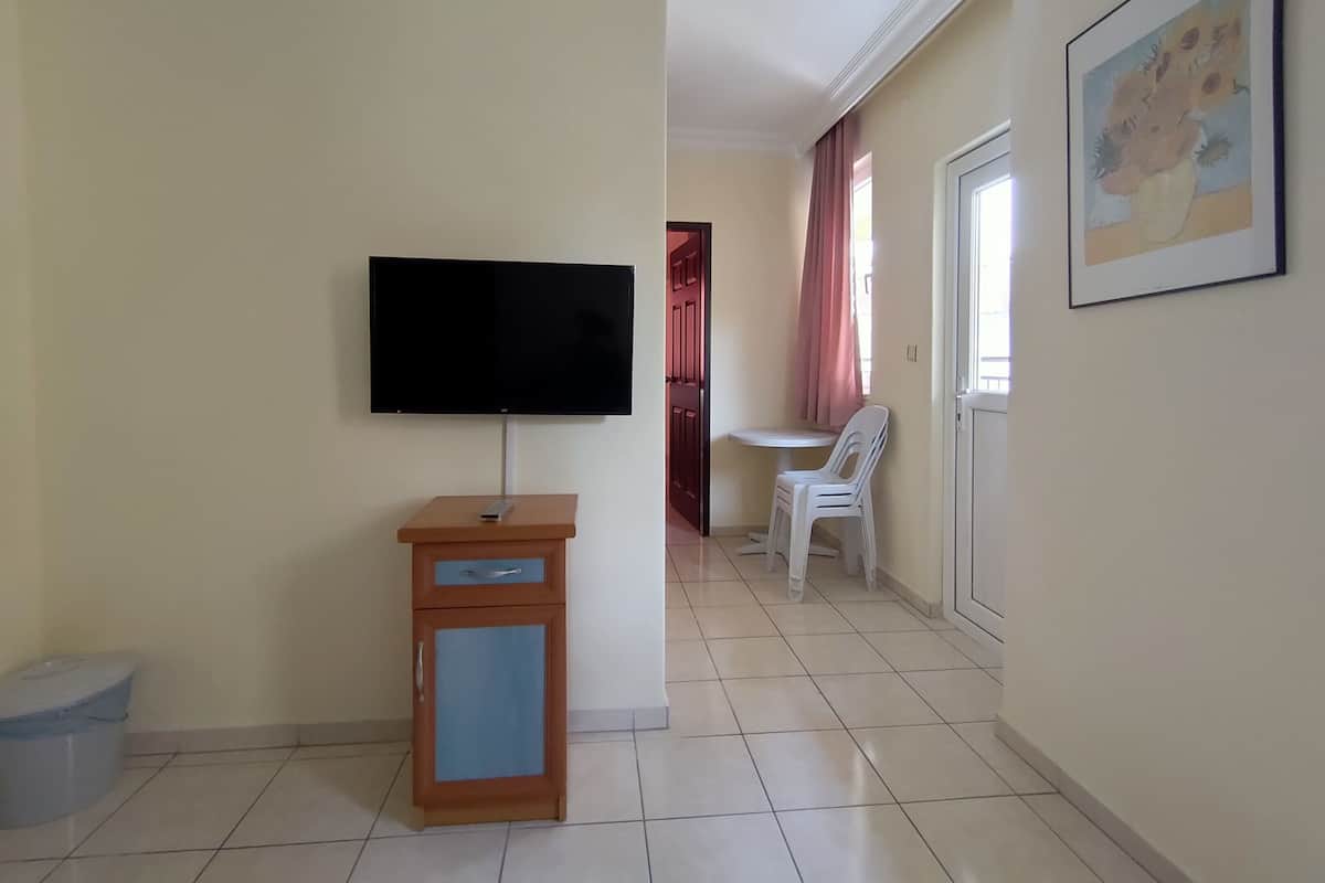 A living room with a flat-screen TV mounted on the wall, a small wooden cabinet with a drawer, a white plastic chair, and a framed picture on the wall.