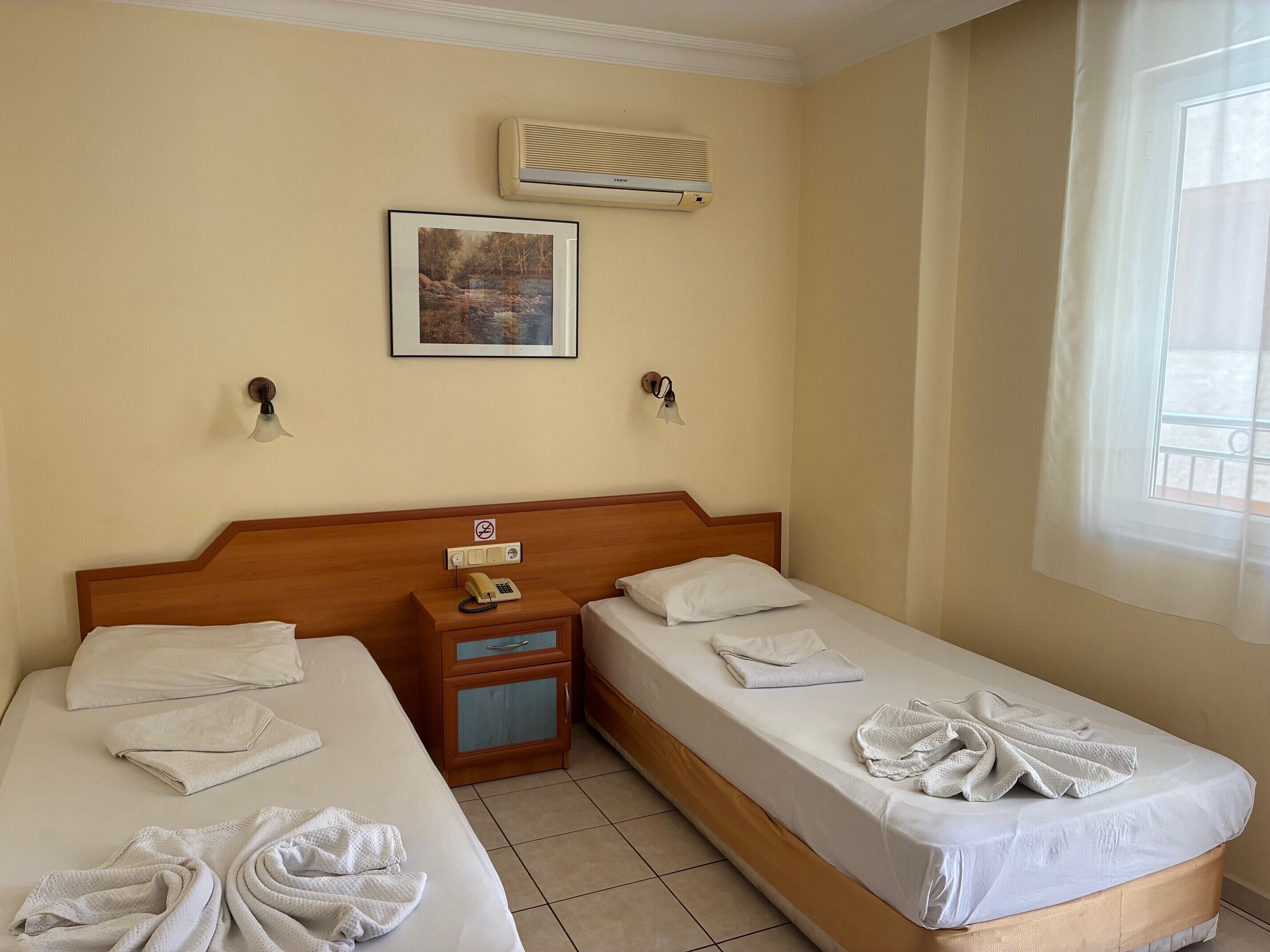 A hotel room with two beds, a wooden headboard, a wall-mounted air conditioner, a framed picture, and a bedside telephone.