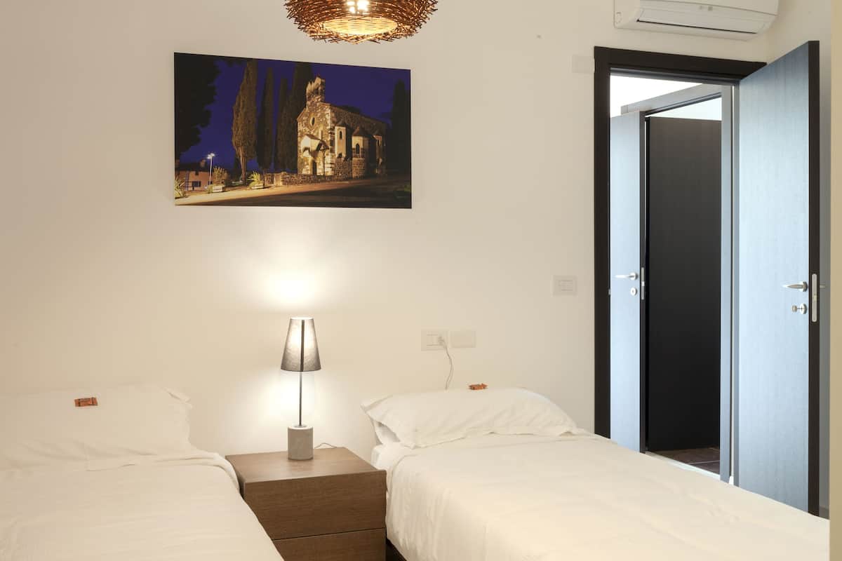 Une chambre avec deux lits, une table de chevet avec une lampe, un tableau au mur et une porte donnant sur une autre pièce.