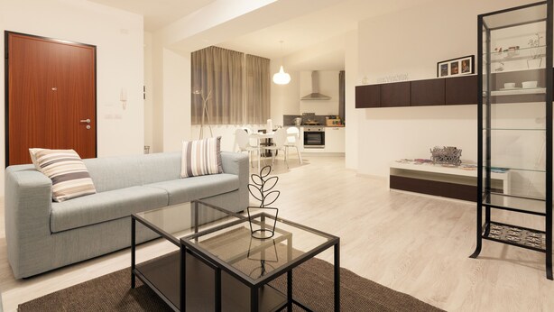 Ein modernes Wohnzimmer mit einem Glastisch, einem grauen Sofa und einer Küche im Hintergrund.