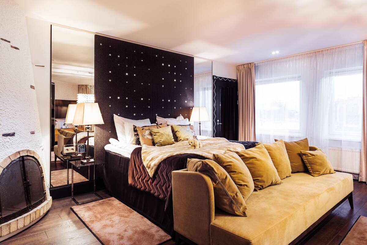 A modern hotel room with a large bed, a sofa, a fireplace, and a view of the city.