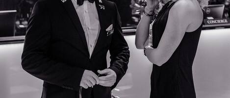 Un hombre con esmoquin y una mujer con vestido posan en un ambiente formal, con suelo a cuadros y un letrero que dice "HOTEL".