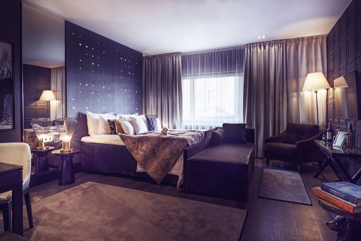 A modern hotel room with a large bed, a sofa, a desk, and a chair. There is a window with curtains, a lamp, and a framed picture on the desk.