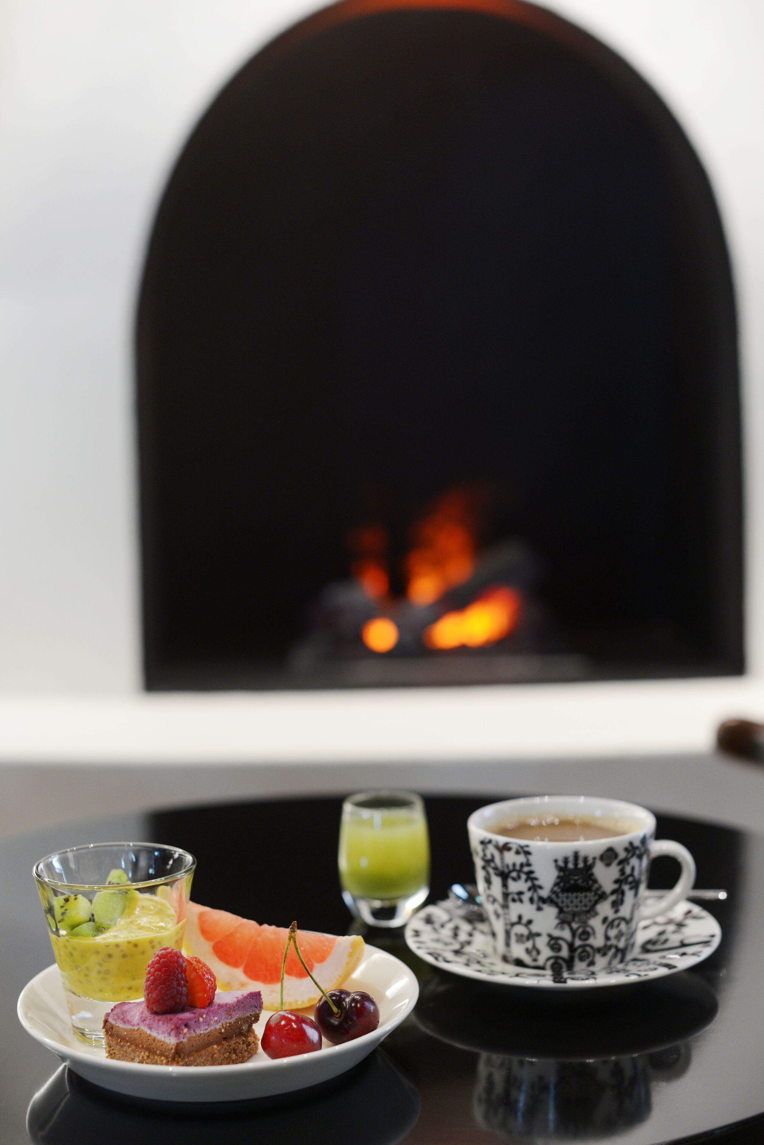 Une table dressée pour le petit-déjeuner, avec une tarte aux fruits, une tasse de café et un verre de jus, devant une cheminée.